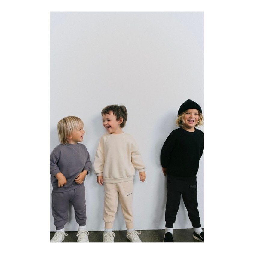 Three young children stand indoors against a white wall wearing casual outfits in gray, beige, and black, smiling and looking at each other.