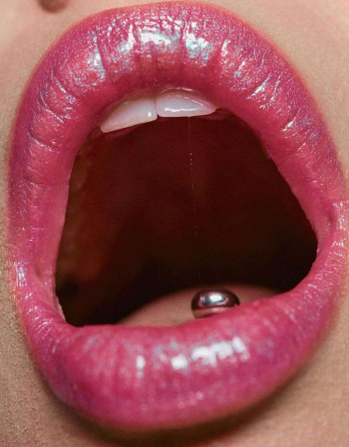 Macro view of a woman's open mouth with glossy pink lipstick, visible teeth, and a silver tongue piercing.