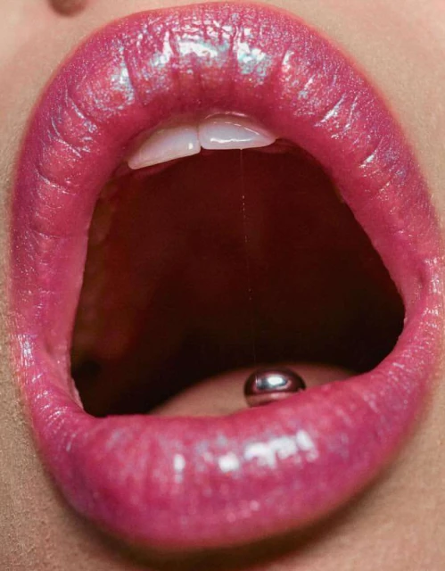 Macro view of a woman's open mouth with glossy pink lipstick, visible teeth, and a silver tongue piercing.