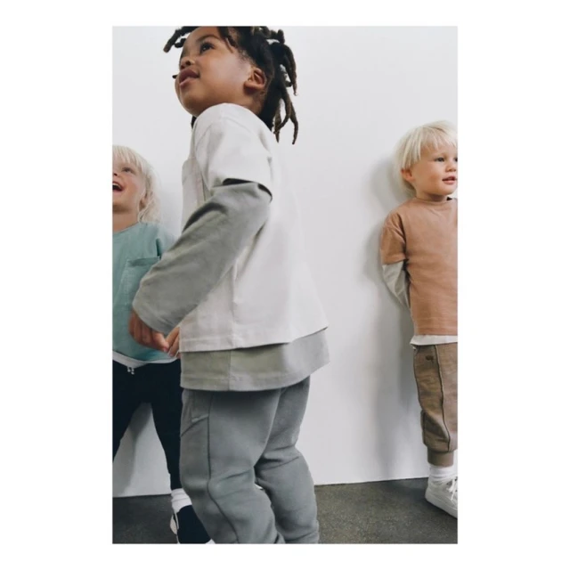 Three young children stand indoors against a plain white wall, wearing casual, layered outfits in neutral colors. One child smiles.