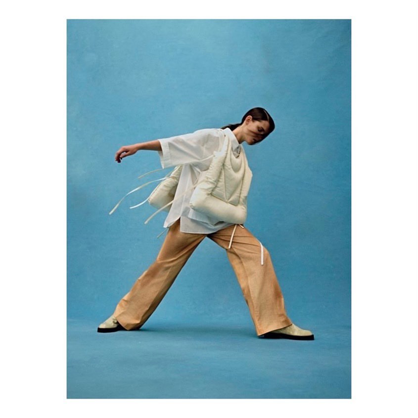 A fashion shot of an adult female model in motion, wearing a white shirt, puffer vest, and beige pants against a light blue background.