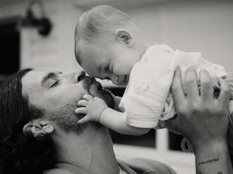 Black and white portrait of a man holding a baby, close together, touching faces. The infant's hands are on the man's face.