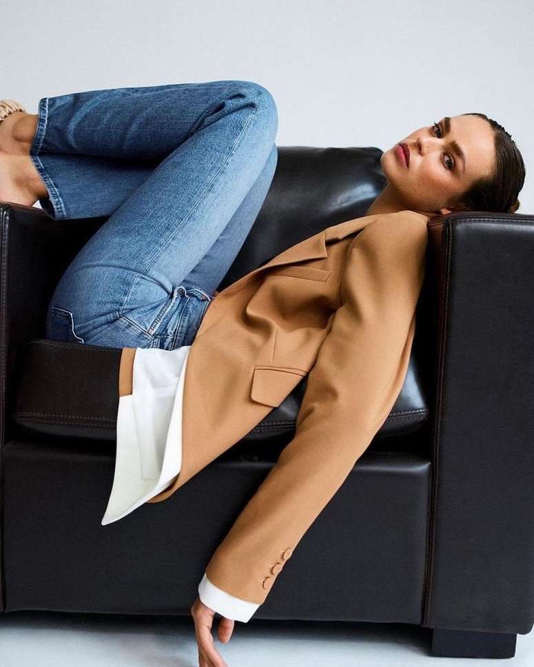 Woman in camel blazer and blue jeans reclining upside down in a black leather armchair against white wall