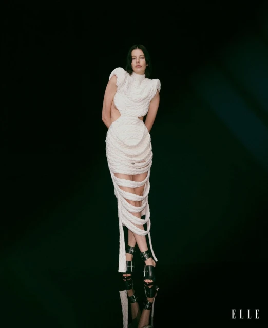 A woman posing in a white, woven dress and black high heels in a fashion studio against a dark background, reflecting on the floor.