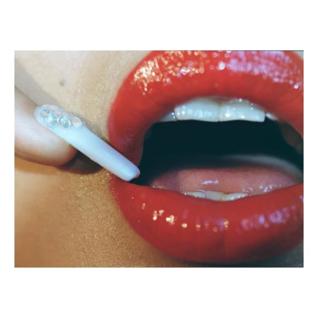 Close-up shot of a person's red lipstick-covered lips with teeth visible, holding a translucent stick with embedded spheres.