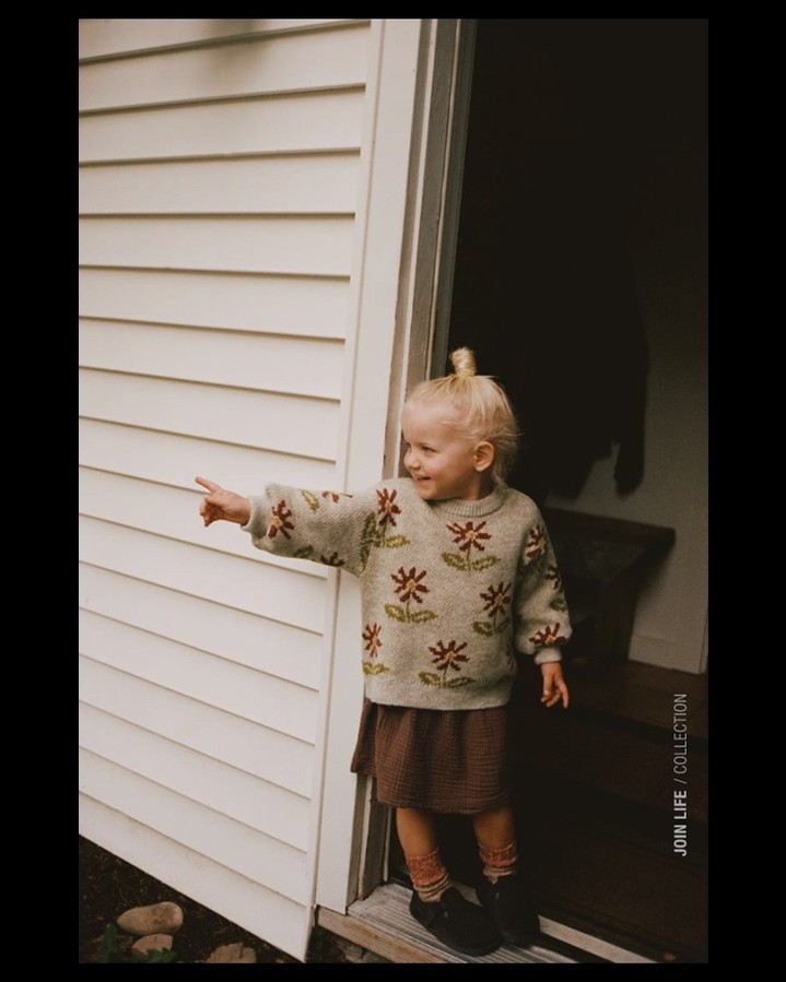A toddler in a flower sweater stands in an open doorway. The young child smiles, looking and pointing toward the left of the frame.