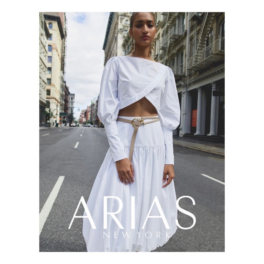 Elegant woman in white top and flowing skirt stands on urban street, buildings line the background, fashion advertising campaign.
