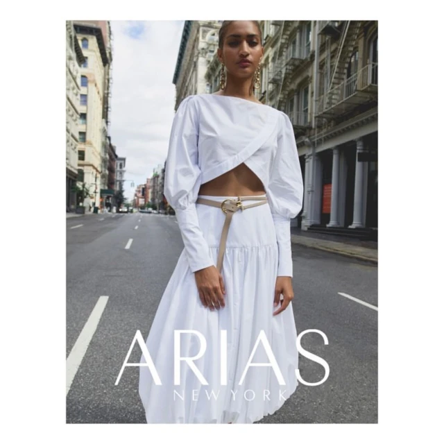Elegant woman in white top and flowing skirt stands on urban street, buildings line the background, fashion advertising campaign.