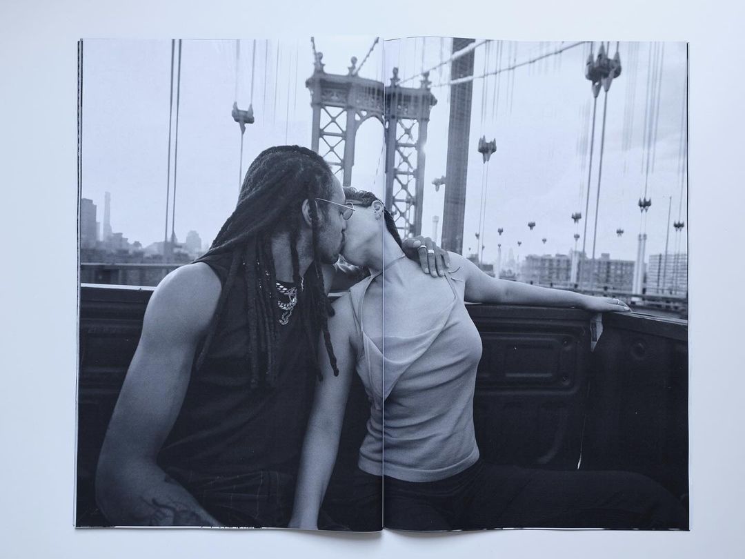 Black and white photograph of a couple kissing in the back of a truck with a bridge in the background and a city skyline.