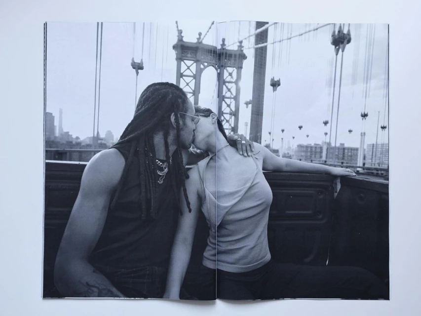Black and white photograph of a couple kissing in the back of a truck with a bridge in the background and a city skyline.