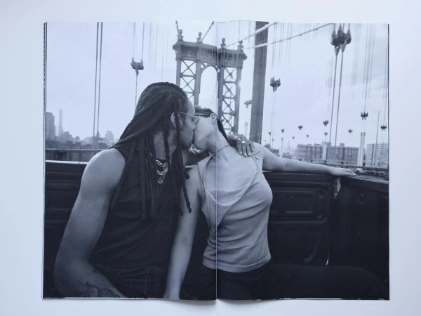 Black and white photograph of a couple kissing in the back of a truck with a bridge in the background and a city skyline.