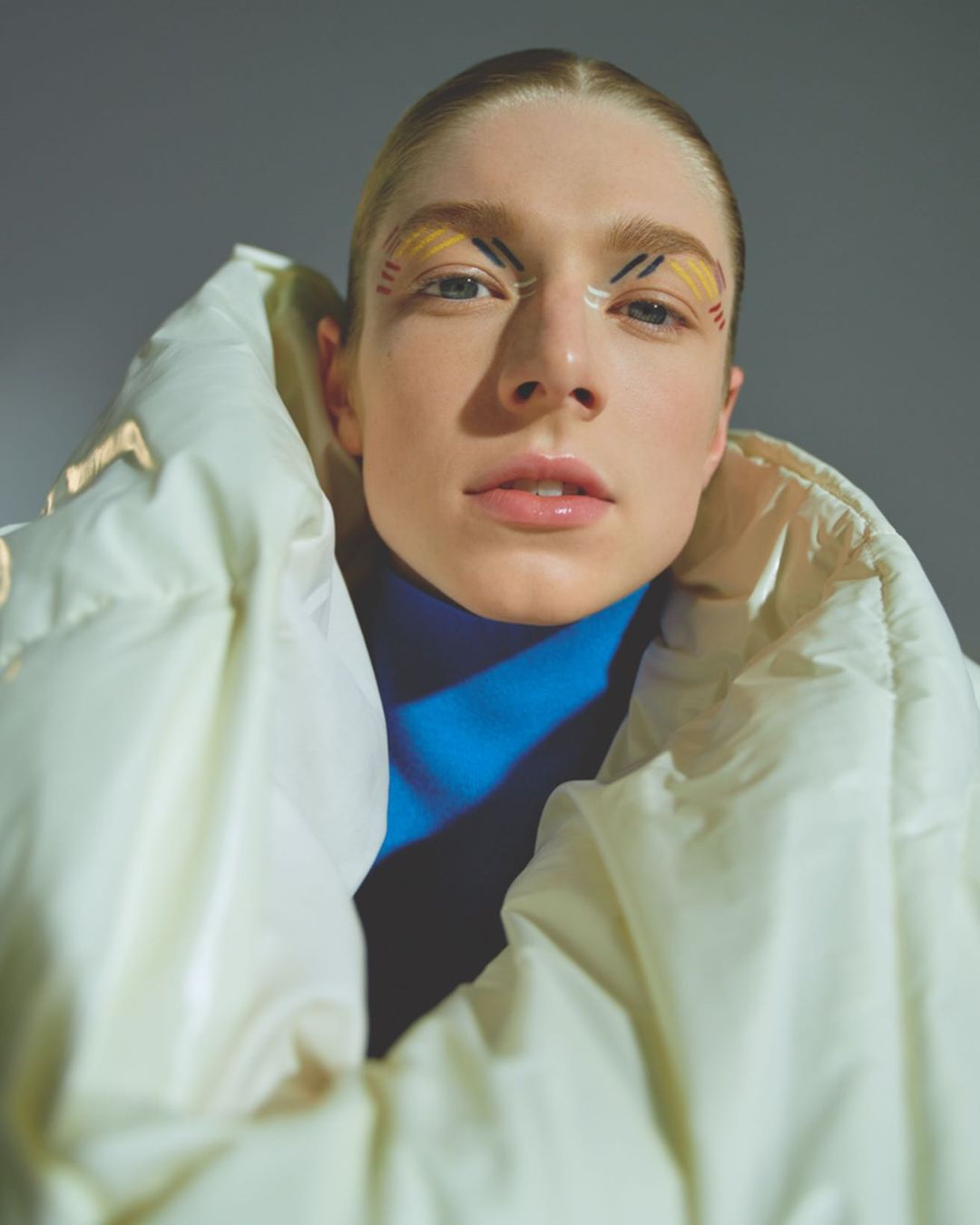 Portrait of a fair-skinned woman with yellow graphic eye makeup wearing a puffy white jacket and blue turtleneck against a gray background.