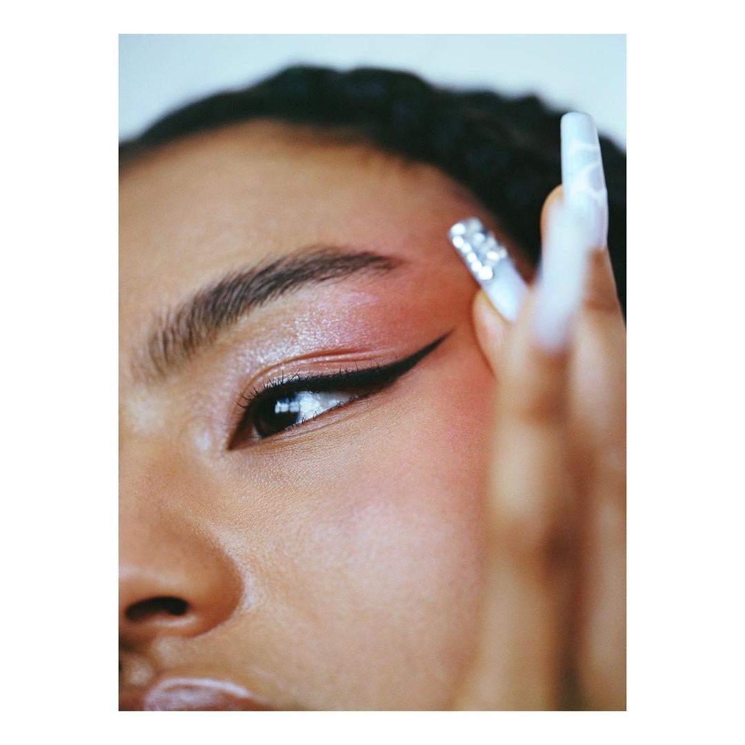 Close up of an adult woman with long jeweled nails and black eyeliner, wearing pink eyeshadow and with fair skin, shot in portrait.
