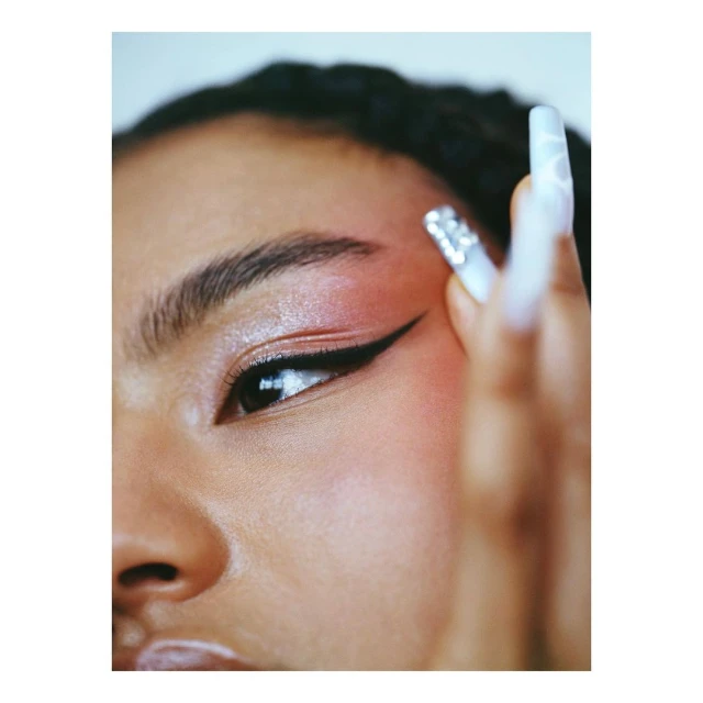 Close up of an adult woman with long jeweled nails and black eyeliner, wearing pink eyeshadow and with fair skin, shot in portrait.