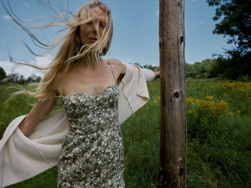 Blonde woman in a floral dress stands by a wooden post in a field of wildflowers wearing a cream cardigan, wind blowing through her hair.