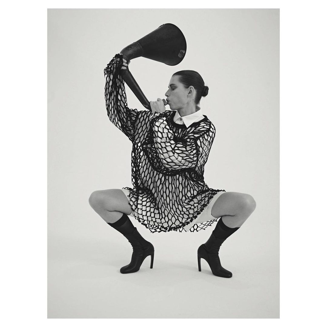 Monochrome studio shot of a crouched adult female with a bun, wearing a mesh top, boots, and holding a large black horn.