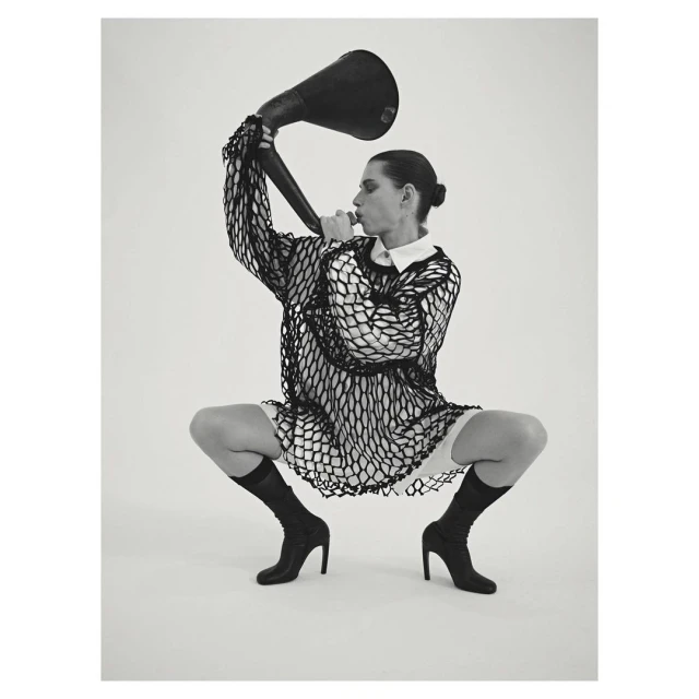 Monochrome studio shot of a crouched adult female with a bun, wearing a mesh top, boots, and holding a large black horn.