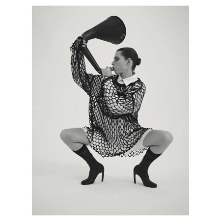 Monochrome studio shot of a crouched adult female with a bun, wearing a mesh top, boots, and holding a large black horn.