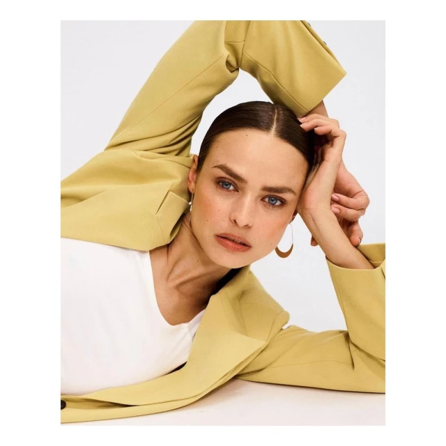 A woman with blue eyes and brown hair, wearing a light-yellow blazer and white top, poses in a studio with arms raised behind head.