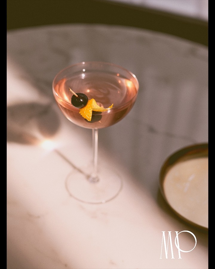 A stem cocktail glass is filled with a pink-hued drink and garnished with a dark cherry and orange peel on a marble table.