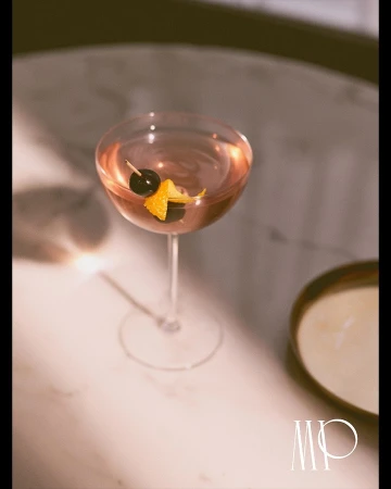 A stem cocktail glass is filled with a pink-hued drink and garnished with a dark cherry and orange peel on a marble table.