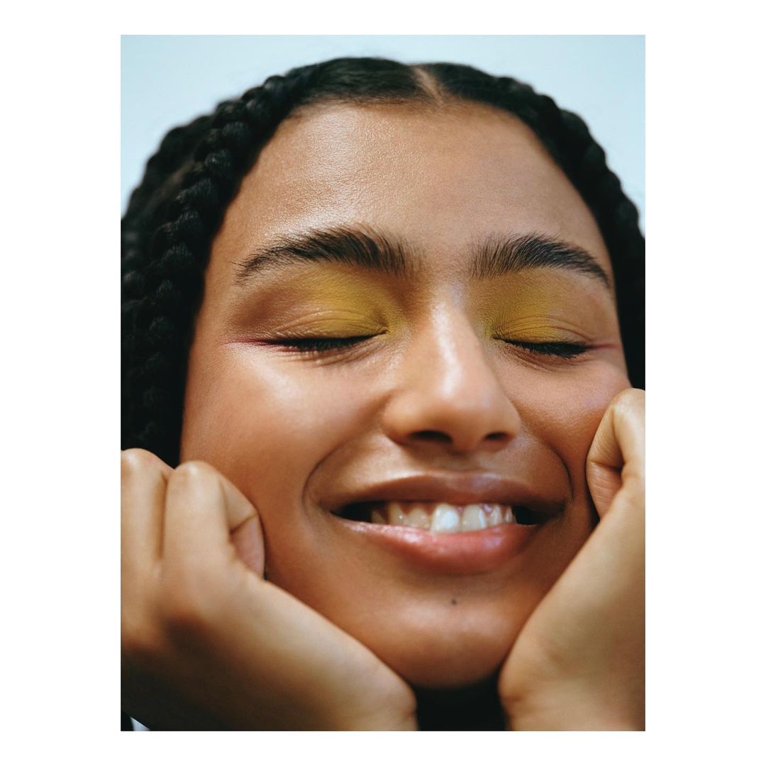 Close-up of a smiling woman with her eyes closed and bright yellow eyeshadow, resting her face in her hands, against a light blue background.