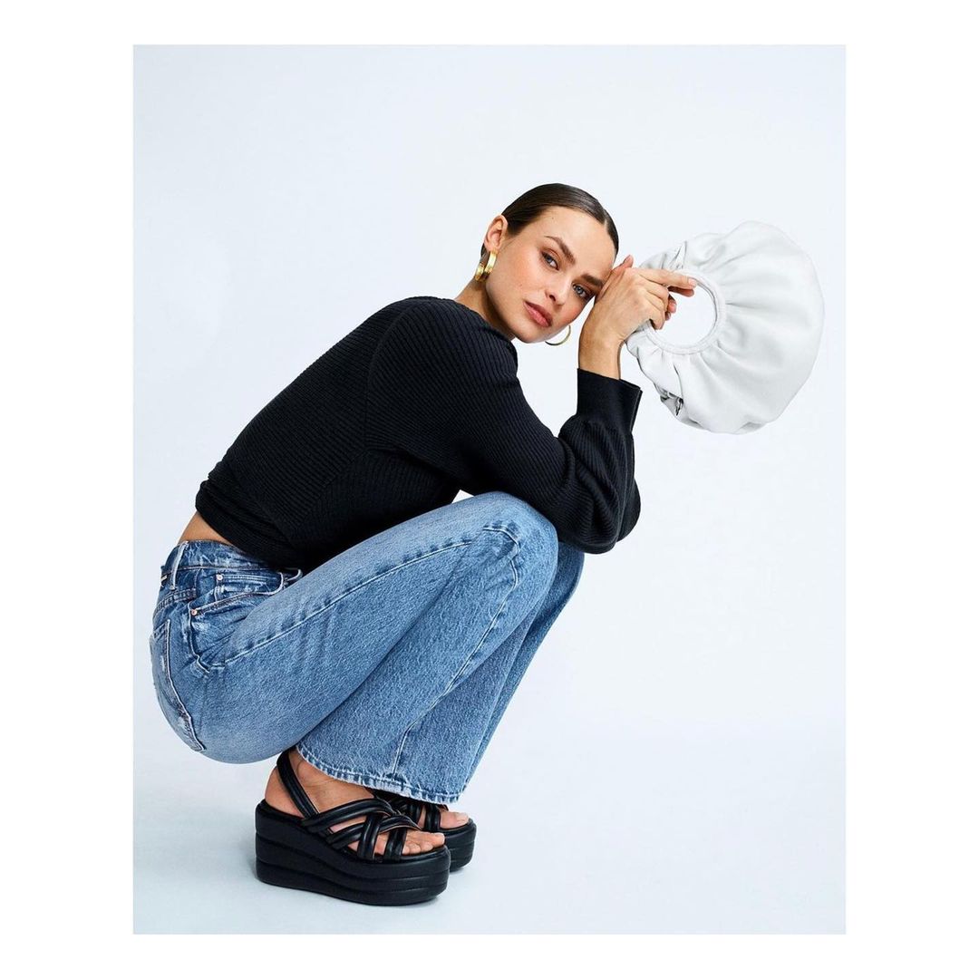 A woman models a black long-sleeve shirt, blue jeans, and black platform sandals while holding a white handbag in a studio setting.