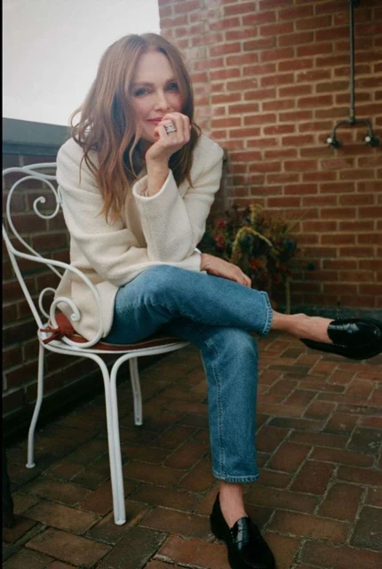 A woman with long auburn hair, wearing a cream-colored jacket and blue jeans, sits on a white chair on a brick patio.