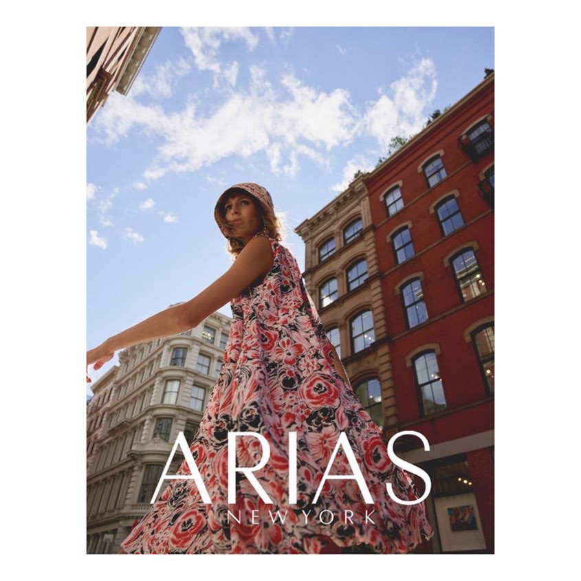 Adult woman in floral print dress and matching bucket hat walking in a city, buildings and blue sky in the background, low angle view.