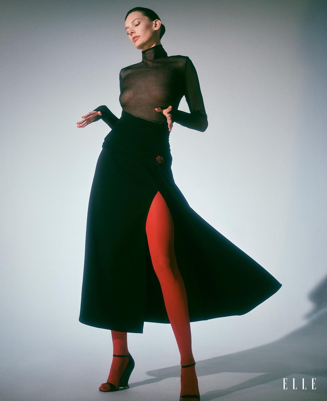 Fashion portrait of an adult woman wearing a sheer top, black skirt with a slit, and red tights against a studio backdrop.