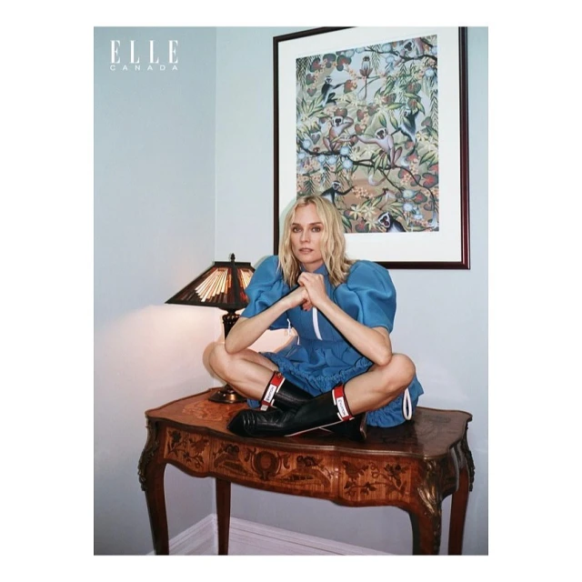 Blonde woman in a blue dress sits cross-legged on a wooden table with an elaborate design, photographed in an indoor setting with natural light.