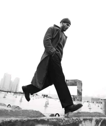 Man wearing a long coat, beanie, and trousers stepping forward in an outdoor urban setting with distant buildings.