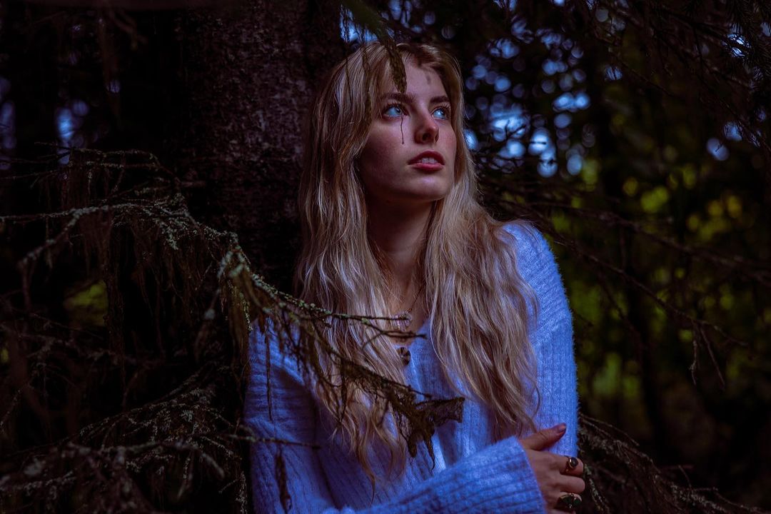 Blonde woman in a blue sweater looks up while standing in a dark forest with a shallow depth of field and bokeh background.