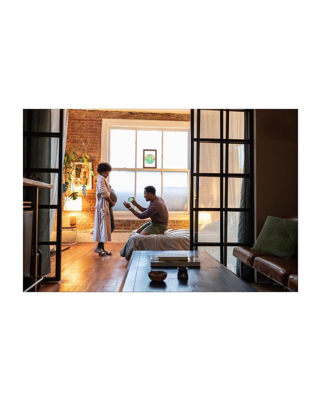 A man kneeling to take a photo of a pregnant woman in an airy apartment with brick walls and wooden floors bathed in warm, natural light.
