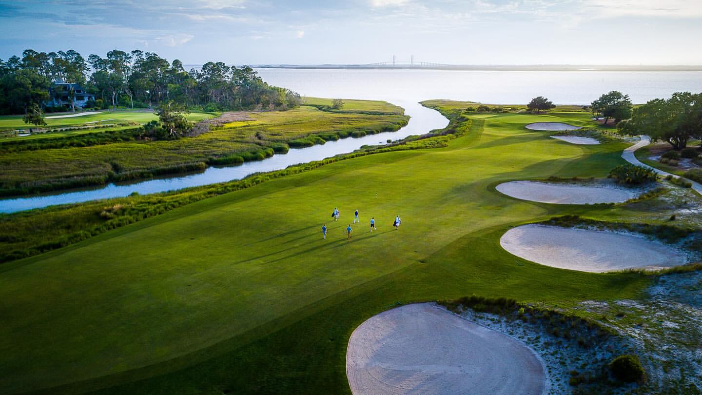 Scenic aerial view of golfers on a lush green golf course beside a winding river leading to the ocean under a partly cloudy sky.