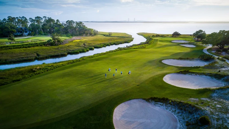 Scenic aerial view of golfers on a lush green golf course beside a winding river leading to the ocean under a partly cloudy sky.