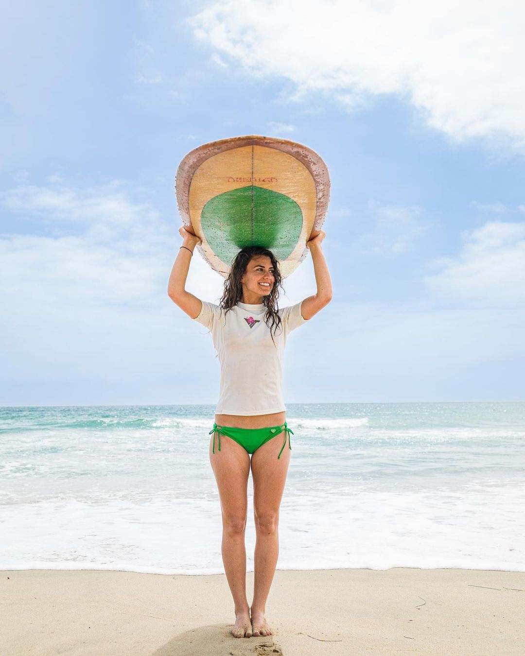 Young woman stands on a beach holding a surfboard over her head, wearing a rash guard and bikini bottom, with a blue sky backdrop.