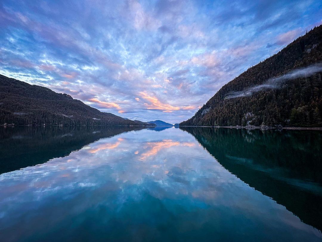 Scenic view of a calm lake reflecting forested mountains under a dramatic sky with pink-tinged clouds at dusk or dawn. Landscape photography.