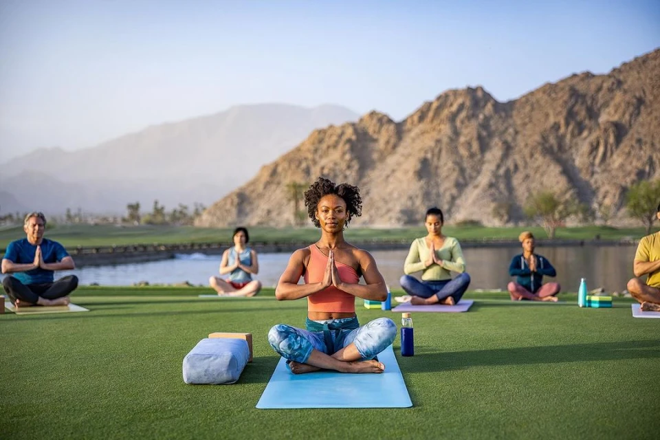 Outdoor yoga class on green lawn with seated adults in lotus pose, hands in prayer position, bottles, yoga mats, mountain backdrop.