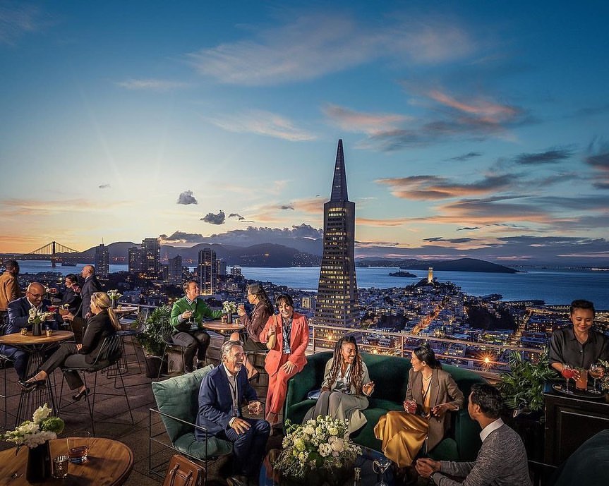 Rooftop gathering at sunset overlooking San Francisco, featuring a group of diverse adults enjoying drinks and conversation.