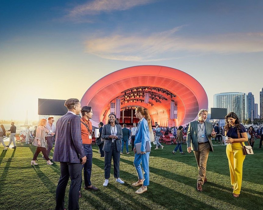 Outdoor corporate event: business people stand on lawn near outdoor stage with backdrop of city buildings on the water at dusk.