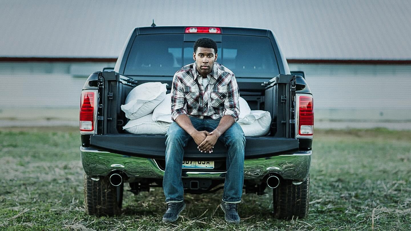 An African-American man sits on the back of a black pickup truck in a rural area, bags are in the truck bed, outdoor setting.