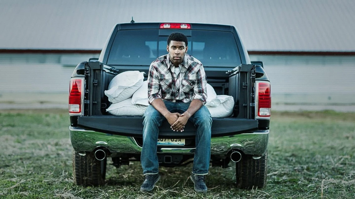 An African-American man sits on the back of a black pickup truck in a rural area, bags are in the truck bed, outdoor setting.