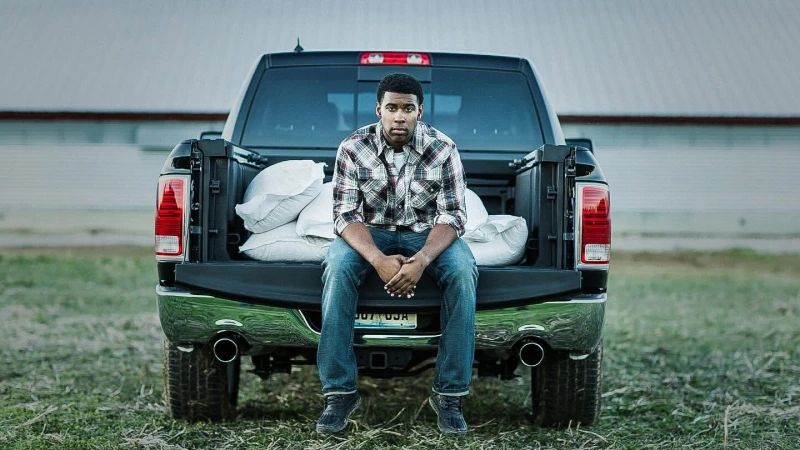 An African-American man sits on the back of a black pickup truck in a rural area, bags are in the truck bed, outdoor setting.