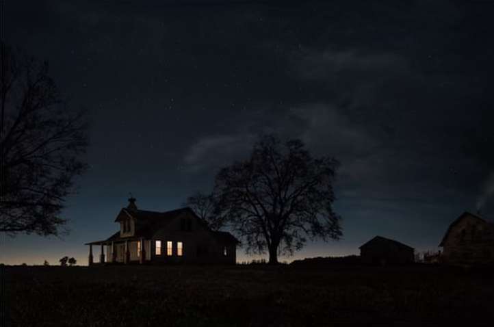 A dimly lit rural night scene shows an old farmhouse silhouette against a starlit sky with scattered clouds and bare trees.