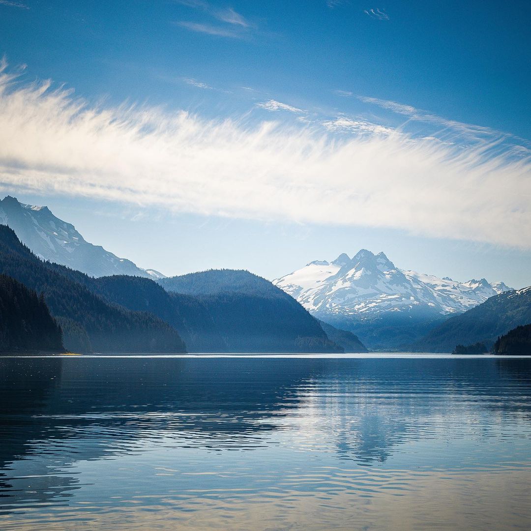 Scenic landscape of a calm lake reflecting forested mountains and snow-capped peaks under a blue sky with feathery clouds.