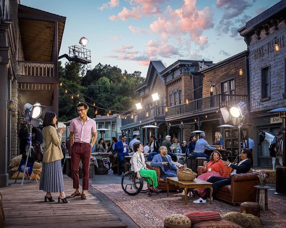 Exterior shot of people gathered in a movie set designed to look like an old western town, production lights visible, warm lighting.