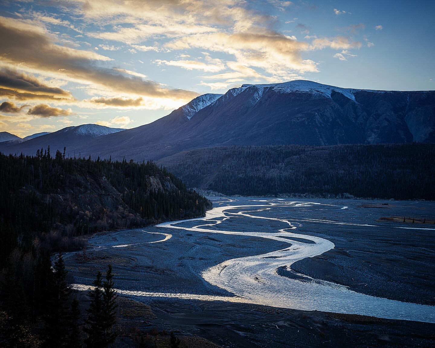 Scenic landscape featuring a winding river flowing through a valley surrounded by dark evergreen trees and snow-capped mountains under a blue sky.