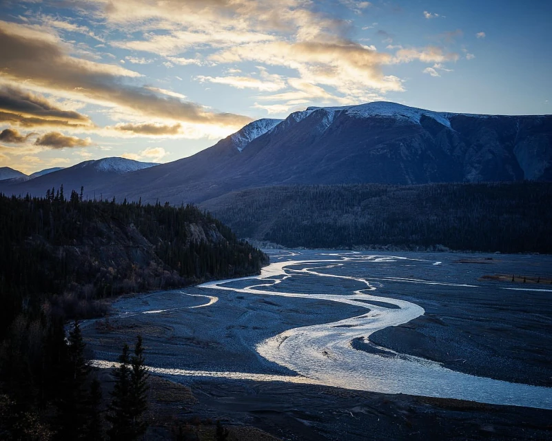 Scenic landscape featuring a winding river flowing through a valley surrounded by dark evergreen trees and snow-capped mountains under a blue sky.