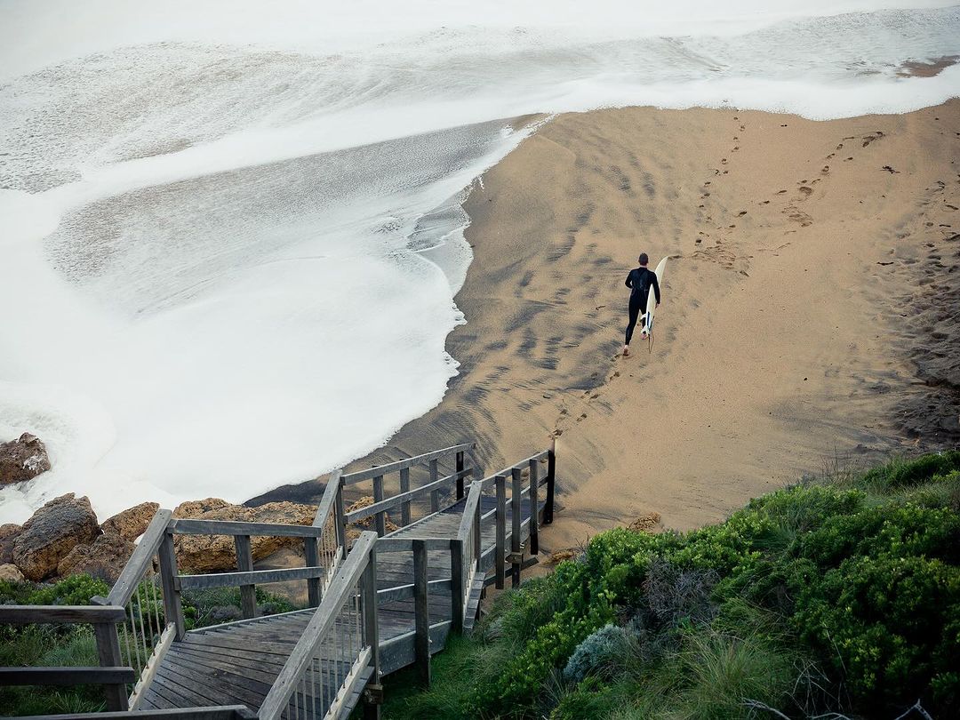 A lone surfer walks along a sandy beach toward the ocean, carrying a surfboard. White waves roll in, meeting the shore, with greenery visible.